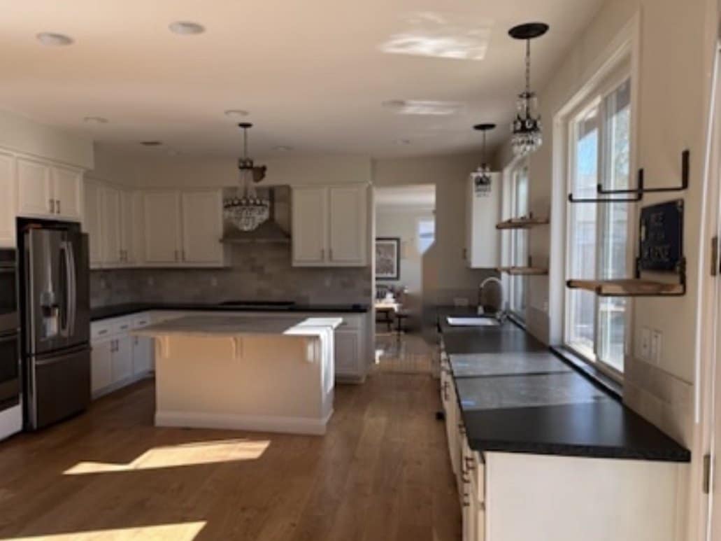 Existing kitchen: white cabinets, black granite countertops, stone-look tile backsplash, stainless double oven and refrigerator, crystal pendants, island with corbels